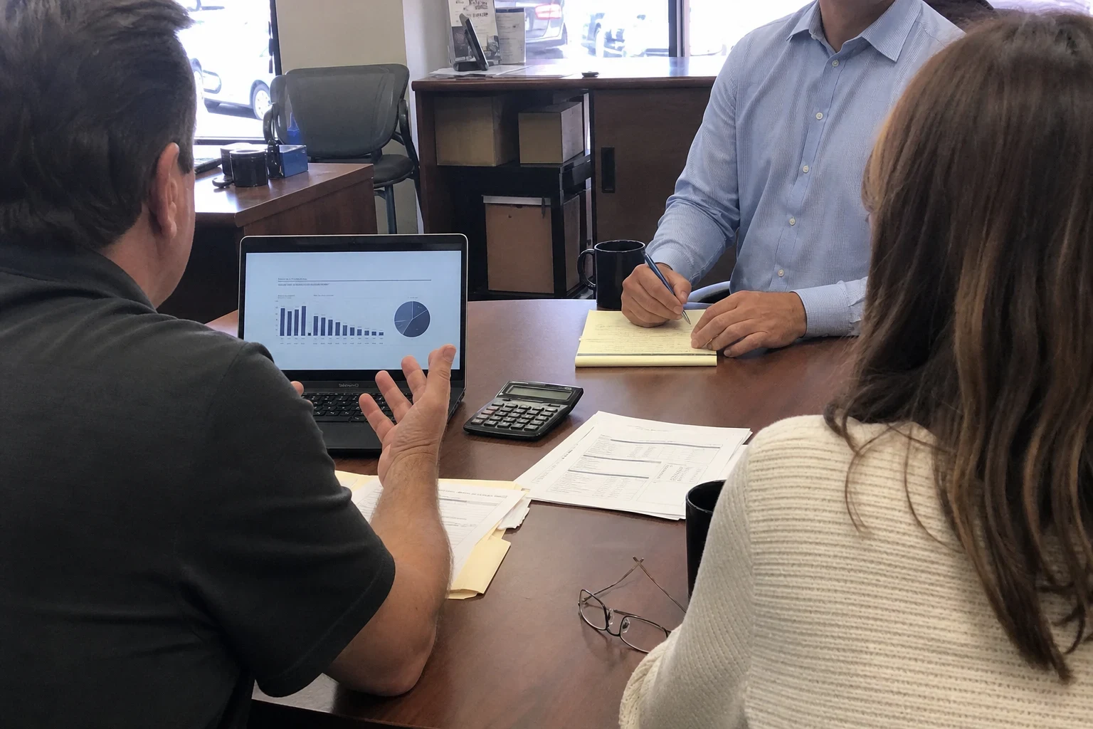 Agent in a blue button-down takes notes on a legal pad while a business owner and his wife review a chart on a laptop at a wooden office table.
