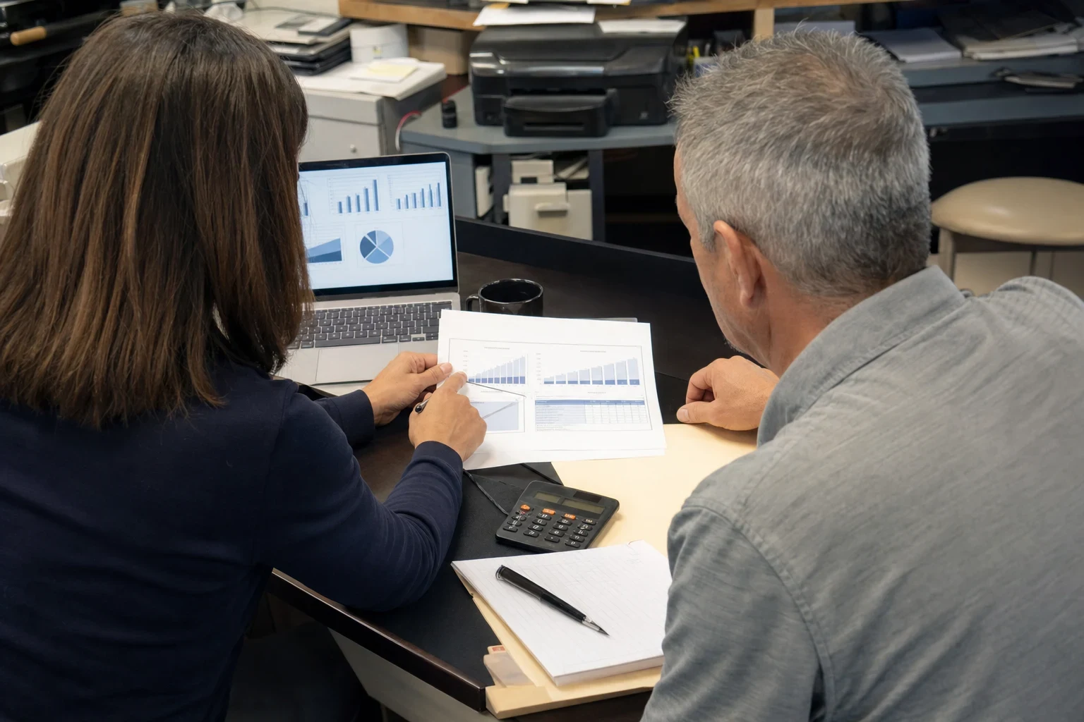 Woman with long brown hair points to printed charts on a desk as a gray-haired man reads along, with a laptop, calculator and notepad in a cluttered back office